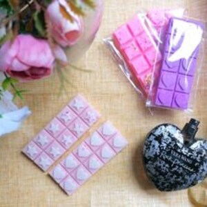 May include: Three wax melts in different colors and scents. The wax melts are in the shape of bars with a grid pattern. The bars are packaged in clear plastic bags. The wax melts are pink, purple, and white.