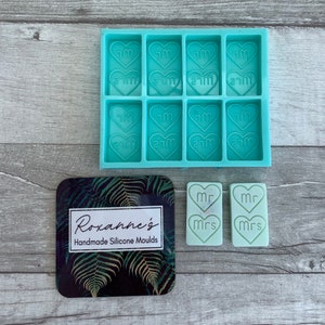 Op de afbeelding: Een turquoise siliconen mal met acht hartvormige holtes, elk met de woorden "Mr" en "Mrs" erop gegraveerd. Twee turquoise hartvormige was smelt met de woorden "Mr" en "Mrs" zijn ook te zien. De mal ligt op een houten oppervlak, en een visitekaartje met de tekst "Roxanne's Handmade Silicone Moulds" is op de achtergrond.