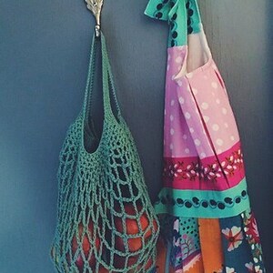 May include: A green crocheted string bag with a variety of red apples inside, hanging on a gold leaf-shaped hook. A colorful patterned apron with pink, white, and turquoise accents hangs on a hook next to the bag.