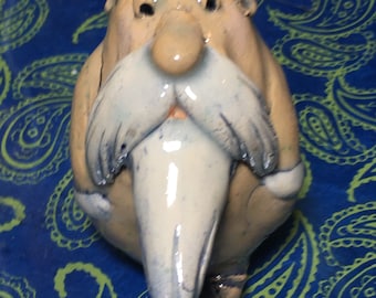May include: A ceramic figurine of a person with a large white beard and a light brown face. The figurine is wearing a white hat and has a large nose. The figurine is sitting on a blue and green patterned surface.