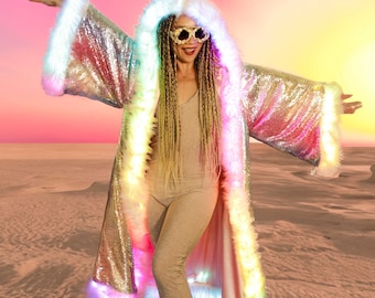 May include: A woman wearing a silver sequined hooded jacket with white fur trim and rainbow LED lights. The jacket has a long, flowing design and is worn over a nude jumpsuit. The woman is standing in a desert setting with a pink and orange sky in the background.