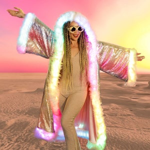 May include: A woman wearing a silver sequined hooded jacket with white fur trim and rainbow LED lights. The jacket has a long, flowing design and is worn over a nude jumpsuit. The woman is standing in a desert setting with a pink and orange sky in the background.