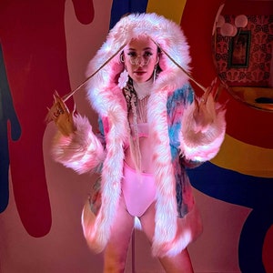 Puede incluir: Una chaqueta de piel sintética de color rosa y azul con estampado tie-dye, con ribete y capucha de piel blanca. La chaqueta se lleva sobre un top de bikini rosa y ropa interior rosa a juego. La modelo lleva botas de plataforma rosa con ribete de piel blanca.