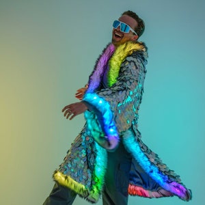 May include: A person wearing a silver sequined jacket with rainbow fur trim and LED lights. The jacket has a long, flowing design and is perfect for a festival or party.