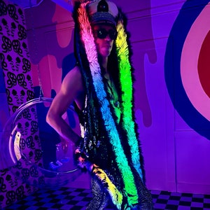 May include: A person wearing a black sequined vest with a rainbow-colored faux fur trim and a matching hood. The person is also wearing black boots and patterned leggings. The background features a checkered floor and a target design.