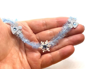May include: A blue and silver garland with snowflake and tree ornaments. The garland is made of tinsel and is perfect for decorating for the winter holidays.