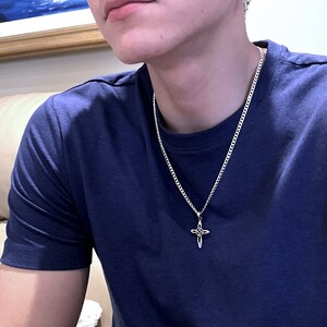 May include: A silver chain necklace with a silver cross pendant. The cross has a decorative design with a circle in the center.