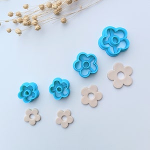 Puede incluir: Vista aérea de cortadores azules con forma de flor y flores de arcilla beige. Los cortadores, de varios tamaños, tienen un diseño detallado. Tallos secos con cápsulas de semillas en la esquina superior izquierda.