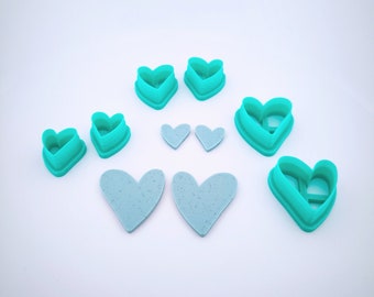 May include: A set of eight teal heart-shaped cookie cutters of various sizes. Two larger teal heart-shaped cookie cutters are in the background, with two smaller teal heart-shaped cookie cutters in the foreground. Two light blue heart-shaped cookies are in the center of the image.