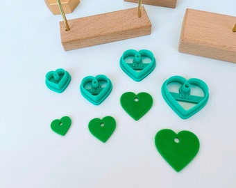 May include: A set of green heart-shaped cookie cutters of various sizes, including a small heart-shaped cookie cutter with the text "62-20" on it, a larger heart-shaped cookie cutter with the text "62-30" on it, and a heart-shaped cookie cutter with the text "62-25" on it. There are also several green heart-shaped earrings on a gold stand.