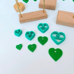Op de afbeelding: Een set groene hartvormige koekjesuitstekers in verschillende maten, waaronder een kleine hartvormige koekjesuitsteker met de tekst "62-20" erop, een grotere hartvormige koekjesuitsteker met de tekst "62-30" erop en een hartvormige koekjesuitsteker met de tekst "62-25" erop. Er zijn ook verschillende groene hartvormige oorbellen op een gouden standaard.