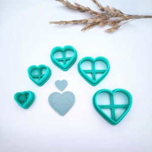 Peut inclure: Ensemble de six emporte-pièces en forme de cœur de couleur turquoise avec un motif en croisillons. Les emporte-pièces sont disposés en dégradé de taille, le plus grand en haut et le plus petit en bas. Un biscuit en forme de cœur turquoise est également inclus dans l'image.