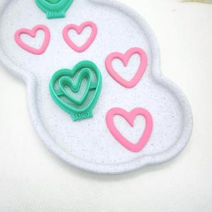 Op de afbeelding: Een verzameling hartvormige koekjessnijders in roze en teal, gerangschikt op een wit, wolkvormig dienblad. De snijders variëren in grootte en ontwerp, sommige met meerdere hartlagen. De lade heeft een gespikkelde textuur.