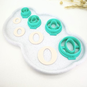 Peut inclure: Ensemble de découpoirs à biscuits circulaires turquoise et découpes de biscuits crème disposés sur un plateau blanc en forme de nuage. Les découpoirs sont numérotés. Des accents floraux séchés sont en arrière-plan.