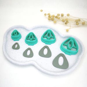 Peut inclure: Ensemble de découpoirs à argile turquoise et gris de différentes tailles, disposés sur un plateau blanc moucheté. Les découpoirs sont triangulaires avec une ouverture ovale. Certains découpoirs sont numérotés.