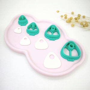 Può includere: Una collezione di formine per biscotti triangolari color turchese con un cilindro centrale, disposte su un vassoio rosa chiaro a forma di nuvola. Sono presenti anche forme triangolari bianche con un foro. Le formine hanno numeri stampati.