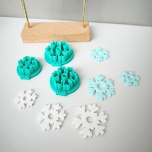 Peut inclure: Boucles d'oreilles blanches en forme de flocon de neige suspendues à un présentoir doré. Des emporte-pièces en forme de flocon de neige turquoise et bleu clair et des découpes de flocons de neige sont disposés autour du présentoir. Thème hivernal.