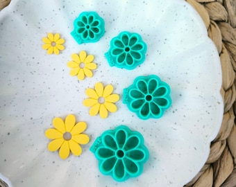 Peut inclure: Un ensemble de six emporte-pièces en forme de fleur. Cinq sont vert turquoise et un est jaune. Les emporte-pièces sont disposés sur une assiette blanche avec une surface mouchetée.