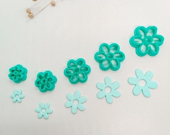 Peut inclure: Un ensemble de sept emporte-pièces en forme de fleur de couleur turquoise de différentes tailles. Les emporte-pièces sont disposés en rangée sur une surface blanche.