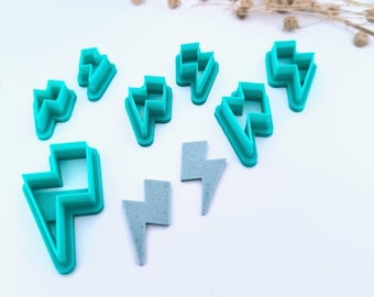 May include: A collection of teal-colored lightning bolt-shaped cookie cutters and two gray lightning bolt-shaped earrings. The cutters are made of plastic and are arranged on a white surface. Dried flowers are in the background.