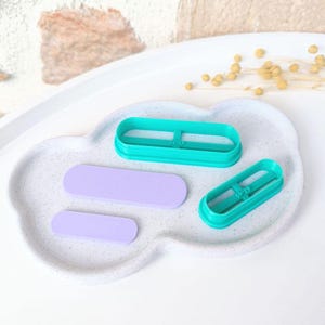 Op de afbeelding: Twee turquoise plastic koekjesvormen in de vorm van rechthoeken met afgeronde hoeken. De vormen liggen op een wit gespikkeld oppervlak met twee paarse fondant rechthoeken.