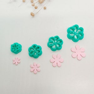 Puede incluir: Conjunto de cinco cortadores de galletas de flores de plástico turquesa y cinco flores de fondant rosa. Los cortadores son de diferentes tamaños y las flores están dispuestas en una fila.