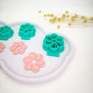 Peut inclure: Des découpoirs à biscuits en forme de fleurs, de couleur turquoise et rose, disposés sur un plateau blanc moucheté. Les découpoirs turquoise portent un numéro. Pour la pâtisserie et l'artisanat.