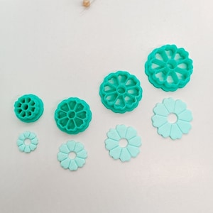 May include: Set of six teal blue plastic flower-shaped cookie cutters. The cutters are arranged in a row from largest to smallest. The largest cutter is a large flower with a heart-shaped center. The smallest cutter is a small, simple flower.