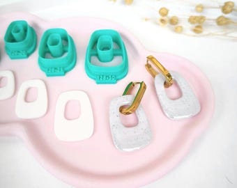 May include: A collection of crafting tools and finished earrings on a pink tray. The tools are teal plastic cutters with the text "128-30". The earrings are white with gold-colored accents. The earrings have a rectangular shape with rounded edges.
