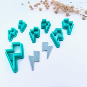 Può includere: Una collezione di formine per biscotti a forma di fulmine color turchese e due orecchini grigi a forma di fulmine. Le formine sono in plastica e sono disposte su una superficie bianca. Fiori secchi sullo sfondo.