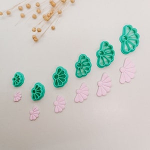 Puede incluir: Un juego de 10 cortadores de galletas de plástico en forma de flor, de color verde y rosa. Los cortadores están dispuestos en una fila del más grande al más pequeño.