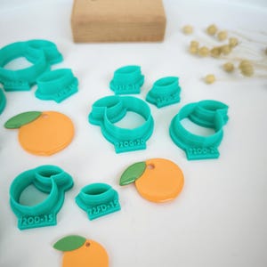 Peut inclure: Un ensemble d'emporte-pièces en plastique turquoise de différentes tailles et formes, notamment des oranges et des feuilles. Les emporte-pièces sont sur une surface blanche avec quelques découpes en pâte d'amande orange.