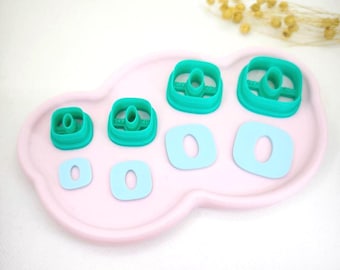 May include: A collection of teal and light blue cookie cutters in various sizes, arranged on a pink, cloud-shaped tray. The cutters are in the shape of a square with a rounded center. The tray is set against a white background.