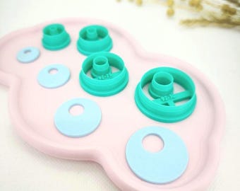 May include: A collection of teal and light blue cookie cutters and cutouts arranged on a pink, cloud-shaped tray. The cutters are various sizes and shapes, including circles and ring shapes. The image is well-lit and shows the items in detail.