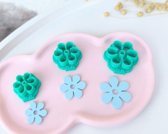 May include: Five cookie cutters on a pink plate. Two are green and shaped like flowers with the text "70-25" and "70-20" on them. Three are light blue and shaped like flowers.