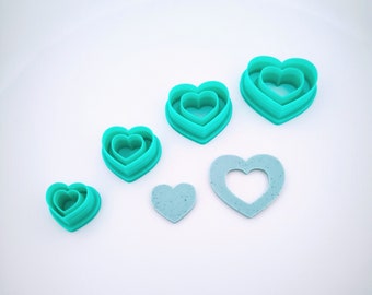May include: A collection of teal heart-shaped cookie cutters in various sizes, alongside two light blue heart shapes. The cutters are nested, showing a layered design. The shapes are arranged on a white surface.