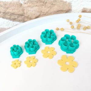 Puede incluir: Un conjunto de cortadores de galletas con forma de flor en varios tamaños y colores. Los cortadores son de color turquesa y amarillo, con marcas de tamaño en los cortadores turquesa. Dispuestos sobre una superficie blanca.