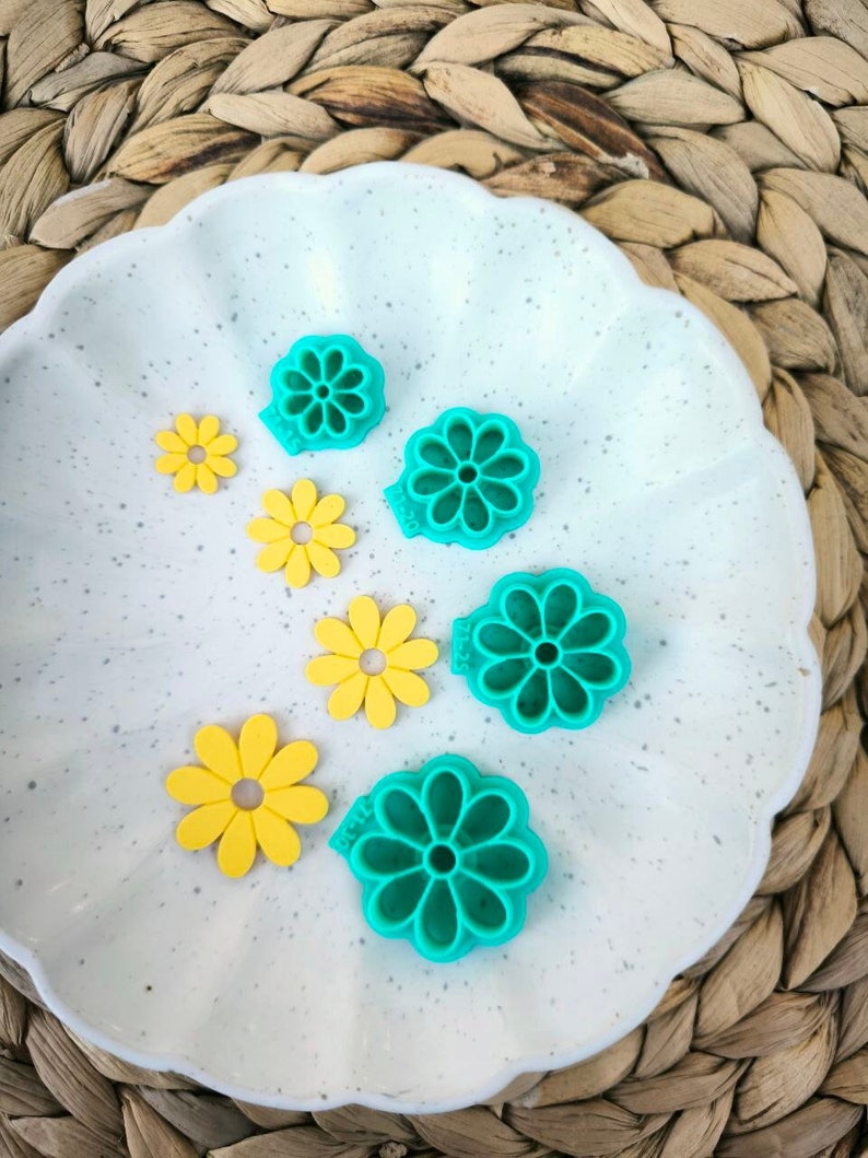 May include: A set of six flower-shaped cookie cutters. Five are teal green and one is yellow. The cutters are arranged on a white plate with a speckled surface.
