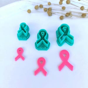 May include: Three teal plastic awareness ribbon cookie cutters with the text 162-20, 162-25, and 162-30 embossed on their bases, accompanied by three corresponding pink awareness ribbon shapes cut out from dough or clay, arranged on a white surface.
