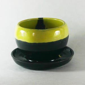 May include: A handmade ceramic cup and saucer set. The cup is a rounded shape, with a bright yellow top half and a black bottom half. The saucer is black. The cup has a black vertical stripe on the inside.