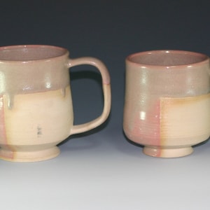 May include: Two ceramic mugs with a light pink and cream glaze. The mugs have a rounded body and a small foot. The larger mug has a handle.