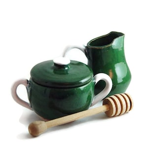 Puede incluir: Un tarro de miel de cerámica verde con tapa y asa blancas, una jarra de crema de cerámica verde a juego y un cucharón de miel de madera.