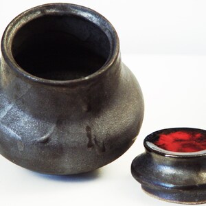 Puede incluir: Un tarro de cerámica negro con tapa. El tarro tiene una boca ancha y un cuerpo redondeado. La tapa tiene un esmalte rojo en el interior.