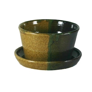 Può includere: Un vaso da fiori in ceramica verde e marrone con un sottovaso abbinato. Il vaso ha una superficie testurizzata e un bordo leggermente svasato.