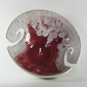 May include: A white ceramic bowl with two handles, decorated with a red and white glaze. The bowl has a unique, abstract design.