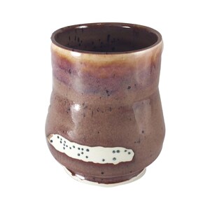 May include: A handmade ceramic mug with a unique, textured surface. The mug features a gradient of colors, transitioning from a light tan at the rim to deep burgundy and brown. A white, oblong accent with dark blue dots adds a distinctive touch to the design.