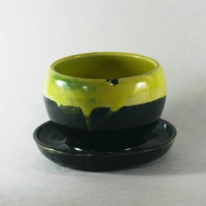 Può includere: Un set di tazza e piattino in ceramica fatto a mano. La tazza ha una forma arrotondata, con una base nera e una parte superiore gialla vibrante che cola sui lati. Il piattino è nero abbinato. Il set è ideale per servire tè o caffè.