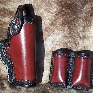 May include: A handcrafted leather holster and magazine pouch set. The holster is burgundy with black trim and decorative stitching. The magazine pouch is also burgundy with black trim. The set is designed for carrying a firearm and extra ammunition.