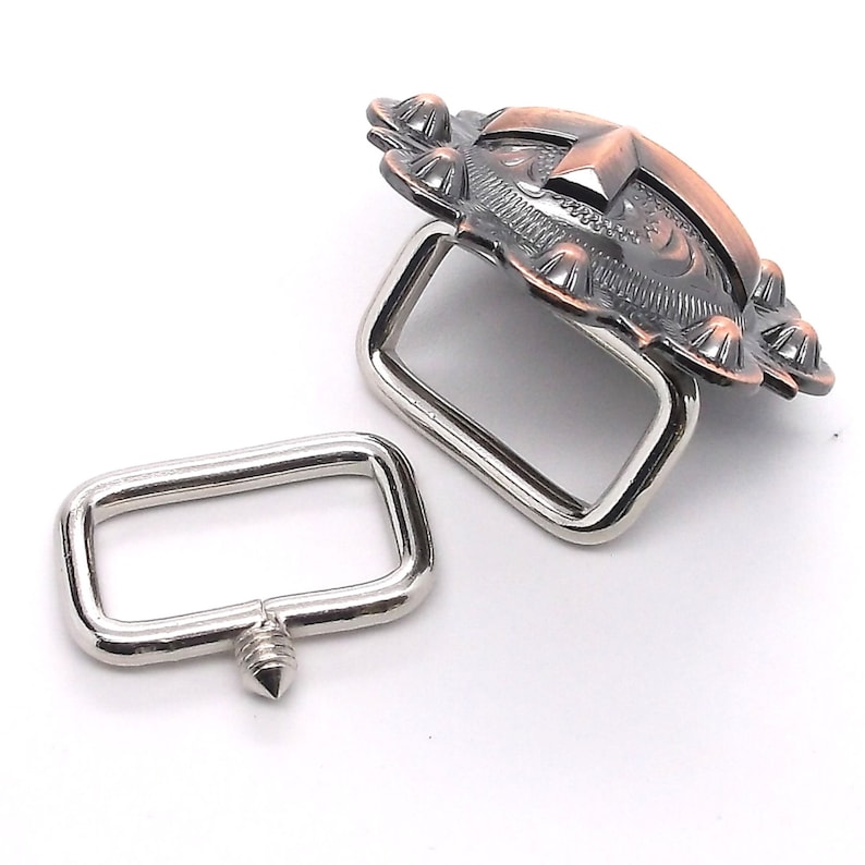 Puede incluir: Una hebilla de metal plateado con un dise&ntilde;o decorativo y ornamentado. La hebilla tiene un acabado de cobre y un patr&oacute;n floral detallado. La hebilla est&aacute; unida a un lazo de metal plateado.
