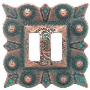 May include: Square, copper-colored metal buckle with two rectangular openings. The buckle has a turquoise patina and decorative floral and geometric patterns. The edges feature a scalloped design with diamond and circular accents.
